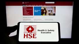 Person holding mobile phone with logo of UK agency Health and Safety Executive (HSE) on screen in front of web page.