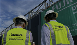 Two people wearing hard hats and yellow Jacobs-branded hi-vis vests in front of a green Fujifilm sign